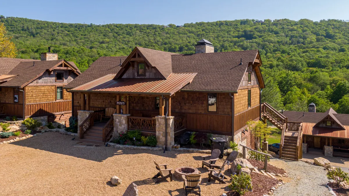 This image features a welcoming wooden porch overlooking a well-defined fire pit area, surrounded by lush greenery. The fire pit is set just beyond the porch, making it easy to imagine transitioning from the deck to fireside evenings. The relaxed atmosphere and clear focus on both the porch and fire pit capture the essence of al fresco leisure and social gatherings under the stars.