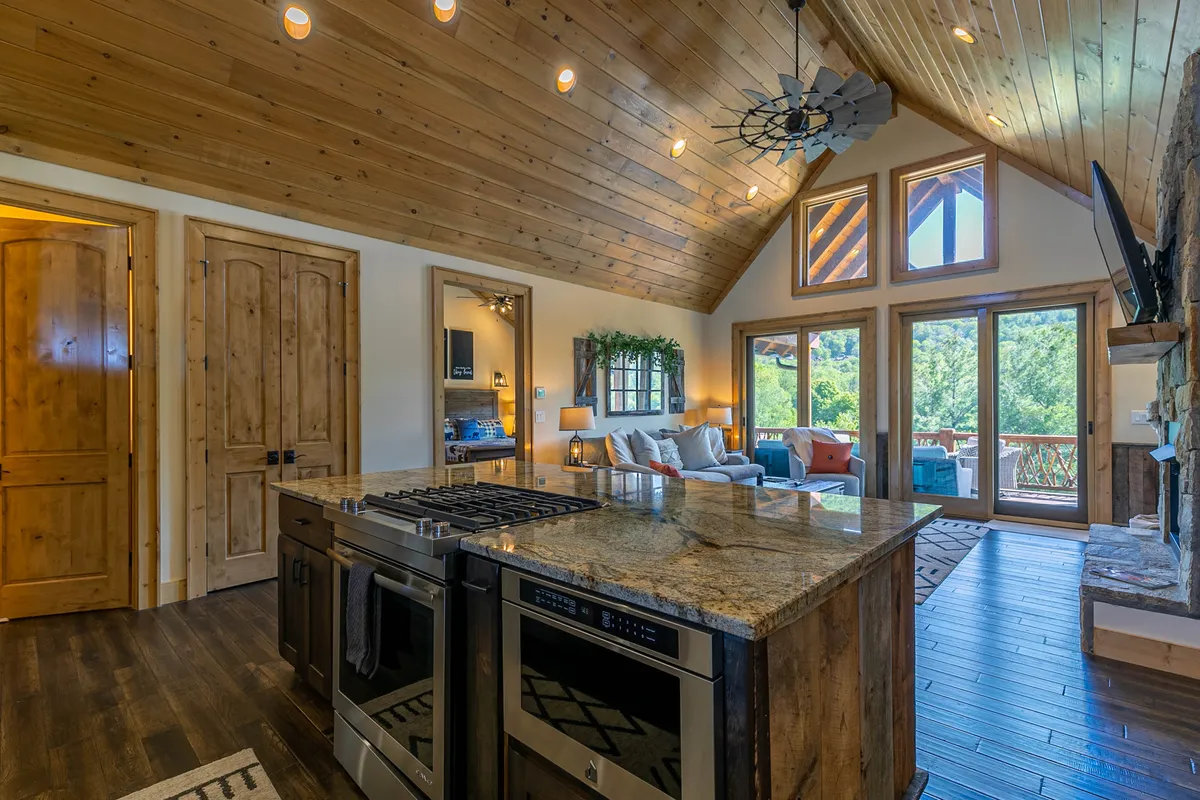 This bright and inviting interior image centers on an open-concept kitchen featuring a granite island, stainless steel appliances, and ample seating. Large windows frame striking mountain views, while the living area offers comfortable seating with colorful pillows and visible wooden ceiling beams. The scene exudes warmth and sociability, ideal for family gatherings or entertaining guests.
