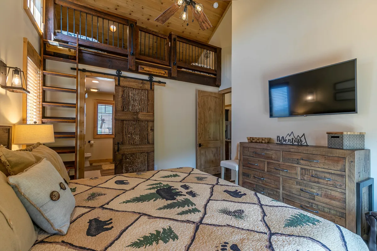 A cozy bedroom with a king bed covered by a patterned quilt, a mounted TV for entertainment, a wooden dresser, and a glimpse of a wooden stairway. The setting is warm and rustic, offering a retreat for relaxation, reading, or streaming shows in comfort.