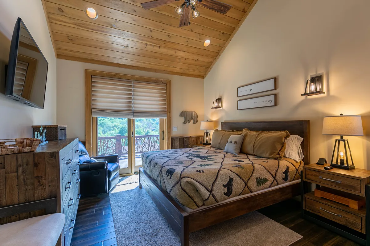 A comfortable bedroom with a king-size bed draped in a patterned blanket, large windows letting in natural light and offering an inviting outdoor view, a wooden dresser, and a ceiling fan overhead. The room is designed to feel cozy and peaceful—ideal for rest and relaxation.