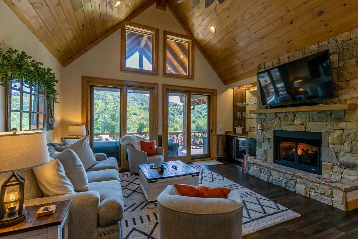 A cozy and stylish living area highlighted by a stone fireplace, plush sofas, a coffee table, and expansive windows offering generous mountain views. The arrangement emphasizes a welcoming atmosphere for relaxing, family gatherings, or enjoying an evening by the fire with a smart TV nearby.