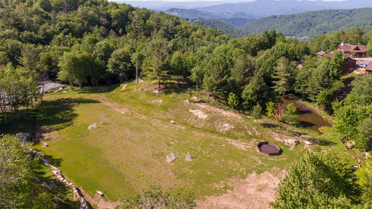 This image showcases a wide open field bordered by trees and with a beautiful mountain view in the distance. There are small structures present, and the scene is bathed in natural light, suggesting the tranquility of nature and the grandeur of the surrounding landscape. The atmosphere is welcoming, with potential for outdoor gatherings and enjoying mountain sunsets.