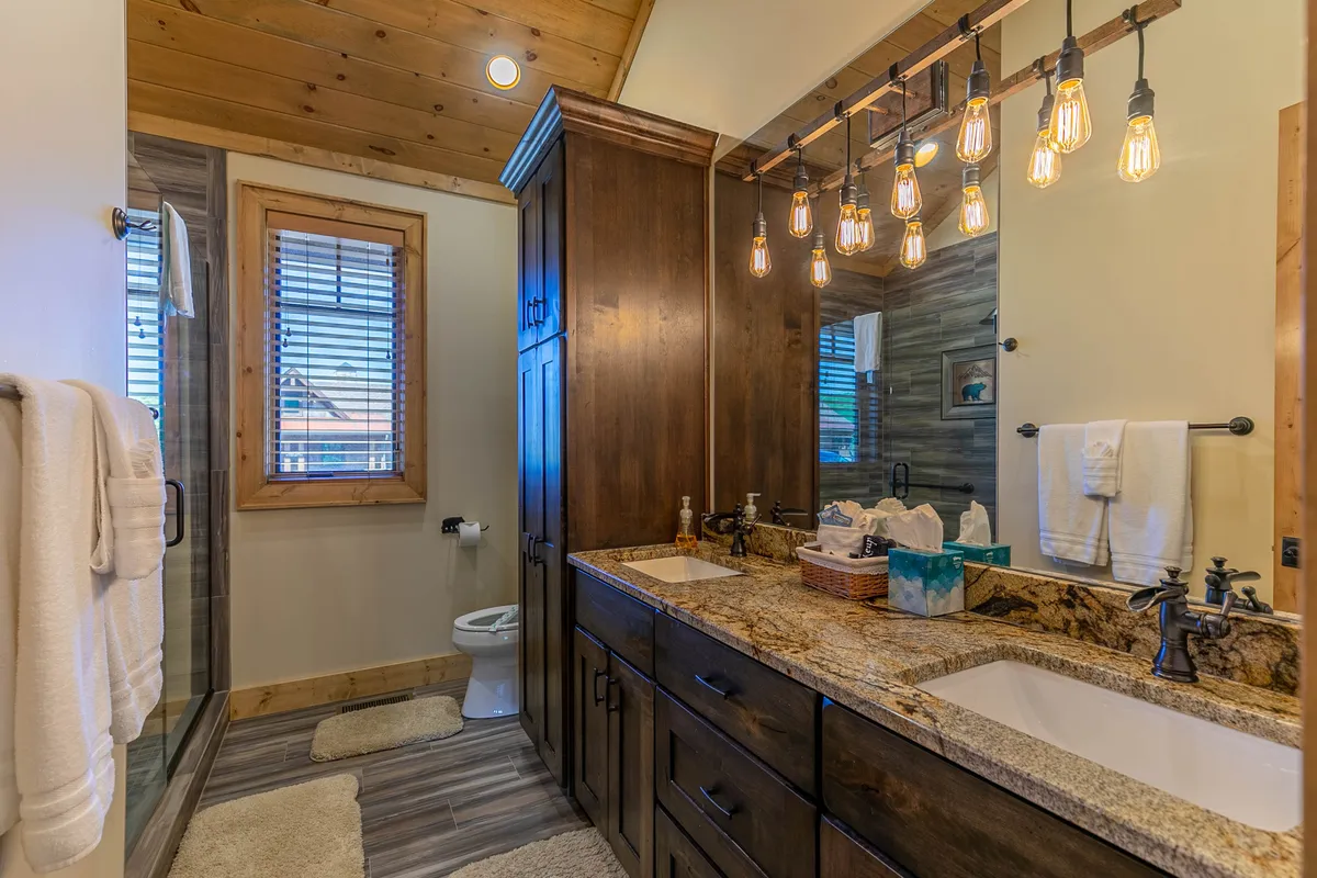 A spacious bathroom featuring a granite countertop, vintage-style light fixtures, wood cabinetry, and a roomy walk-in shower. The calm, comfortable mood suggests a peaceful retreat for self-care routines.