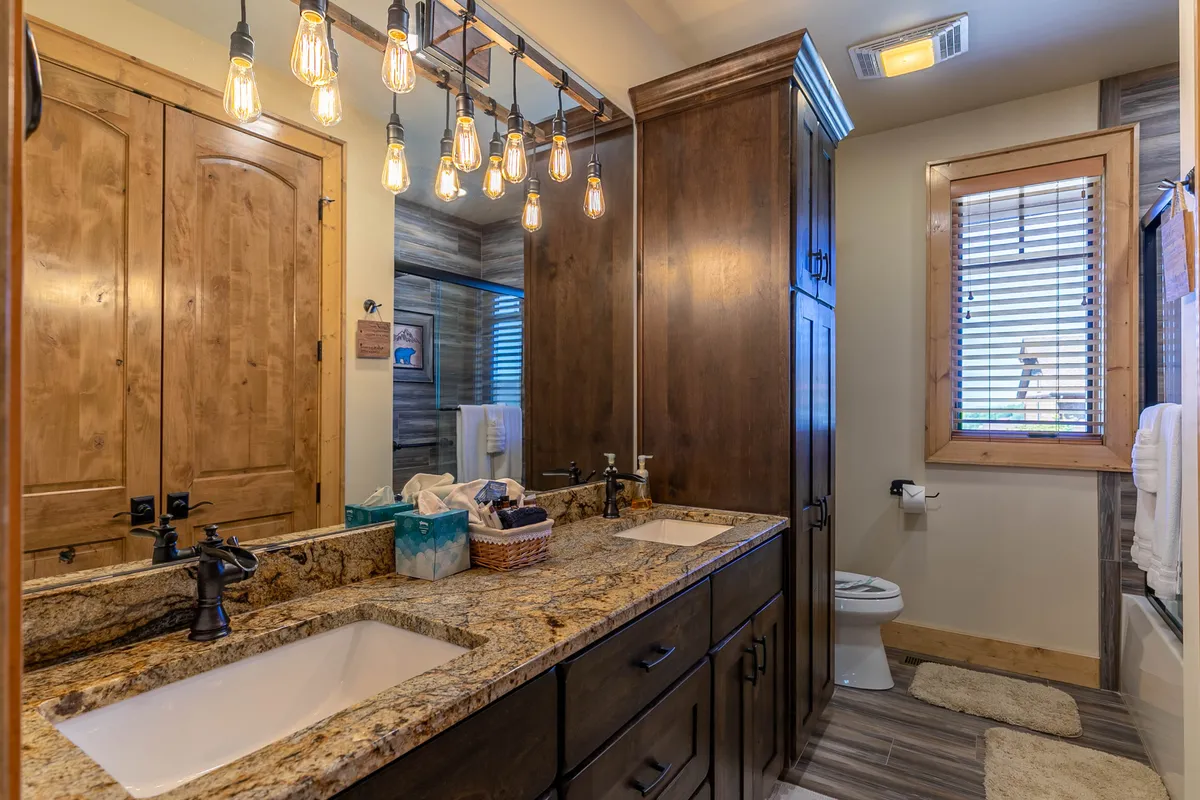 A modern bathroom showcasing a double sink granite vanity with hanging light fixtures and wooden cabinets below, complemented by a framed window that lets in natural light. The layout and finishes evoke a spa-like, relaxing ambiance.