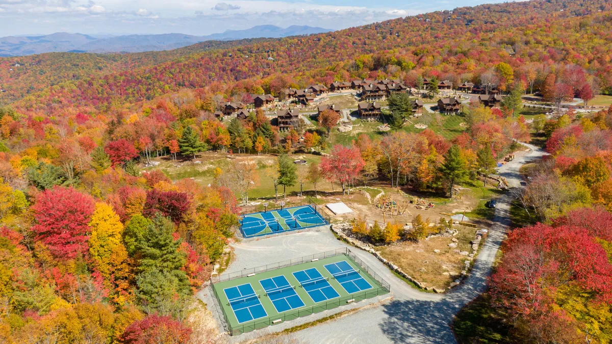 This image captures colorful autumn foliage surrounding tennis courts and a mountain landscape dotted with cabins. The scene is vibrant and energetic, with clear visual cues of outdoor recreation and the striking beauty of the Blue Ridge Mountains in fall.