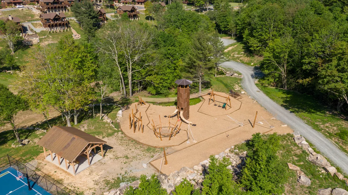 This image features a vibrant playground structure surrounded by trees, a basketball court, and a winding pathway. Families and children can be seen enjoying the outdoors, with plenty of open space for play and relaxation.