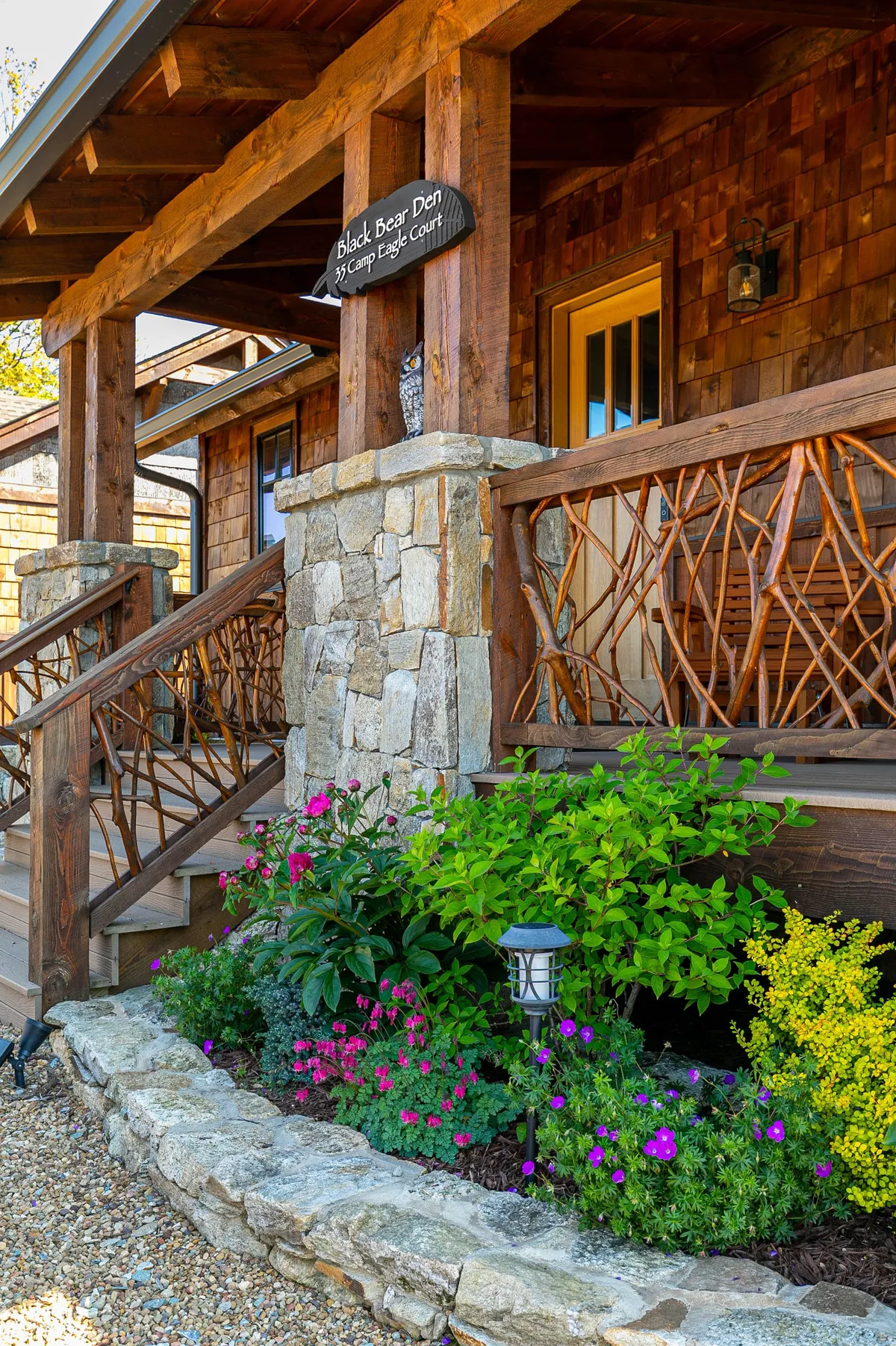 This inviting exterior shot features stone steps leading to a porch with a wooden railing, surrounded by colorful flowers. The house sign is visible, and the mood is warmly welcoming, suggesting a cozy return after a day of wine tasting or exploration.