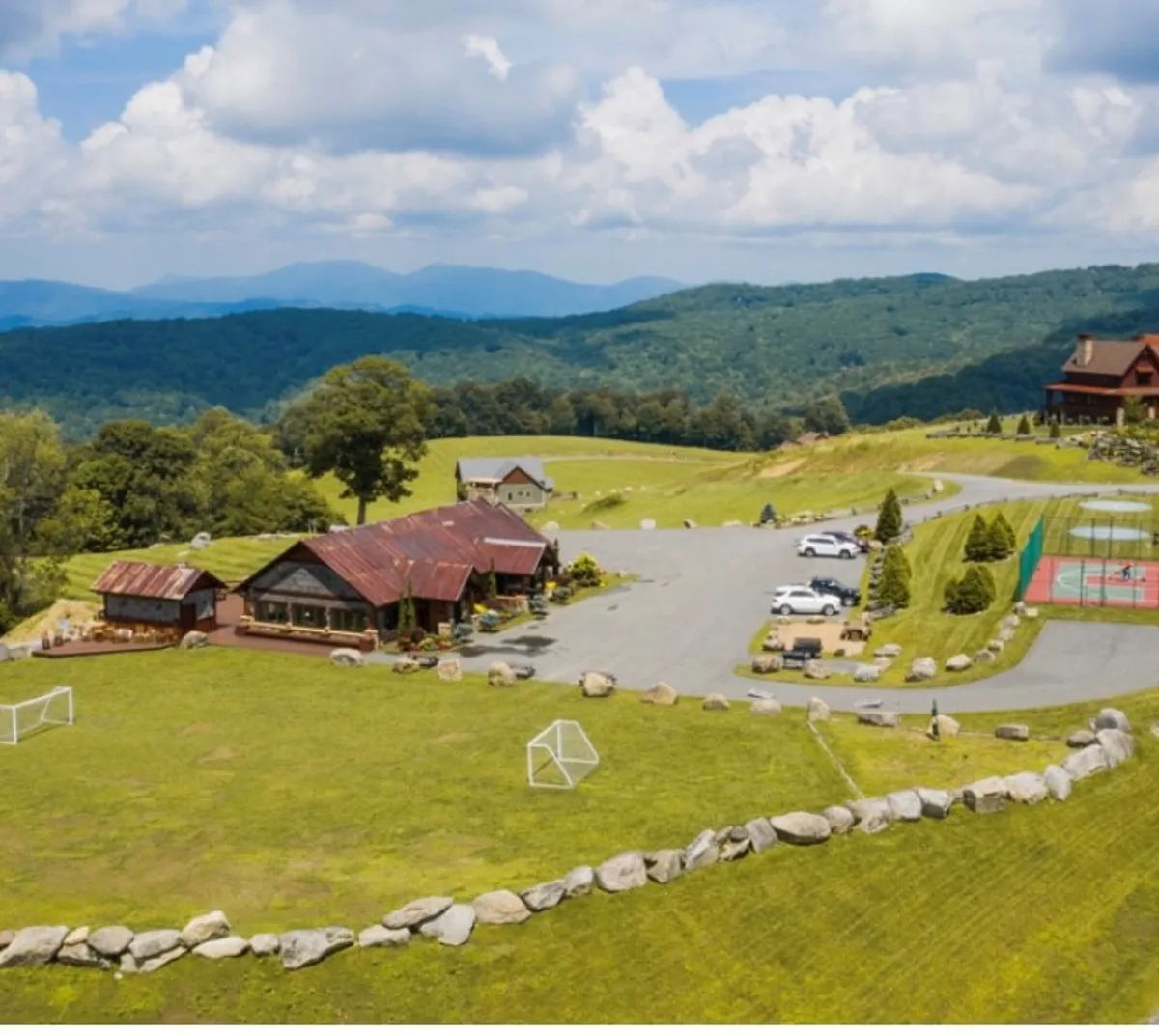 A wide exterior shot of a rustic lodge with soccer goals, a tennis court, a mountain backdrop, and a clearly visible parking area. The setting highlights open recreational spaces and convenient parking for guests.