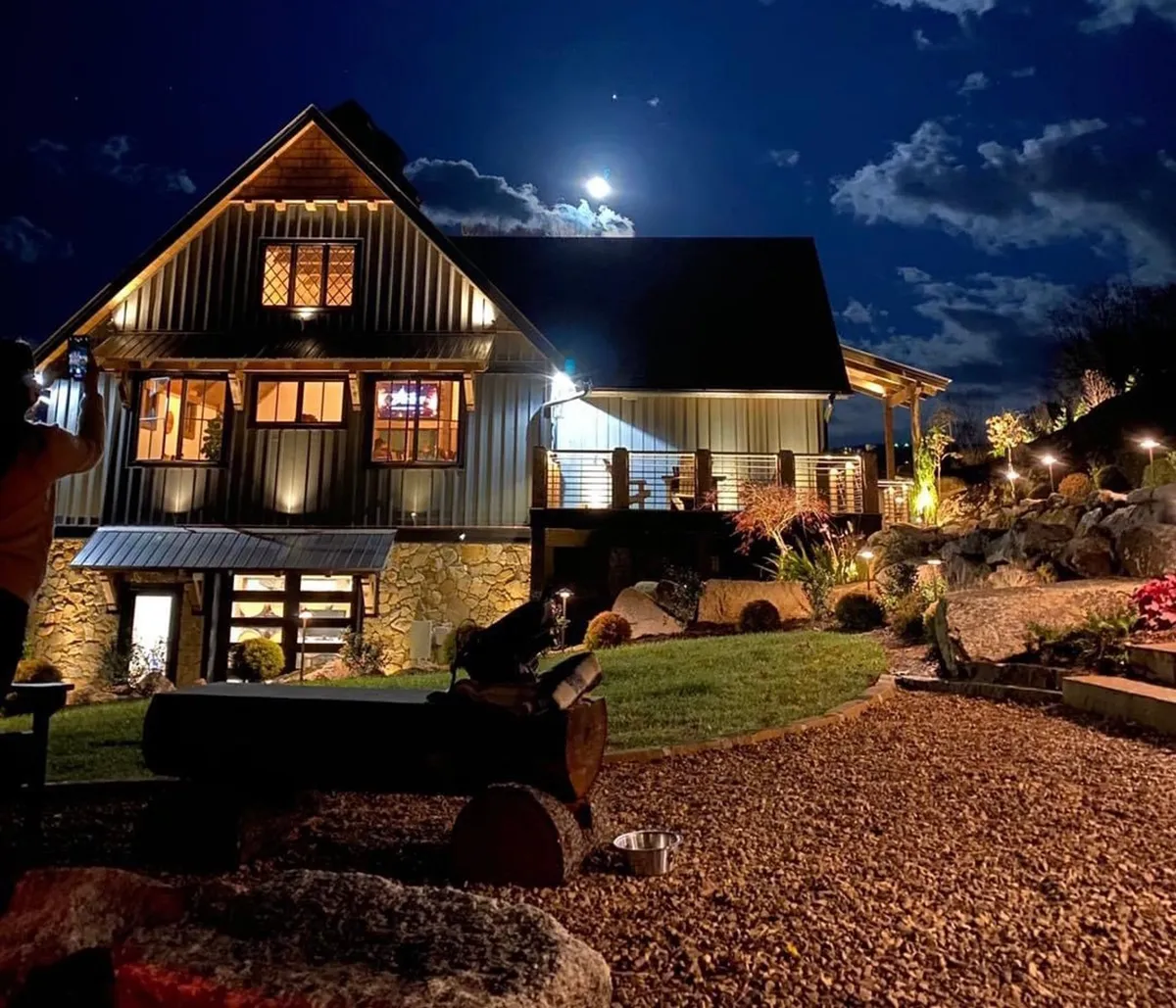 This exterior night scene highlights a well-lit house with a landscaped yard, featuring a glowing fire pit in use and the moon shining above. The ambiance is cozy and peaceful, reflecting the inviting mood of stargazing and fireside relaxation described in the section. The visible fire pit and outdoor space reinforce evening outdoor activities.
